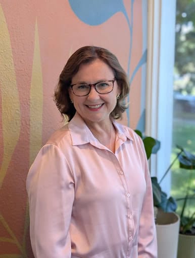 A woman with shoulder-length brown hair and glasses smiles while standing indoors. She wears a light pink blouse and stands in front of a colorful wall with plant designs, embodying hope and positivity on her journey of recovery from eating disorders.