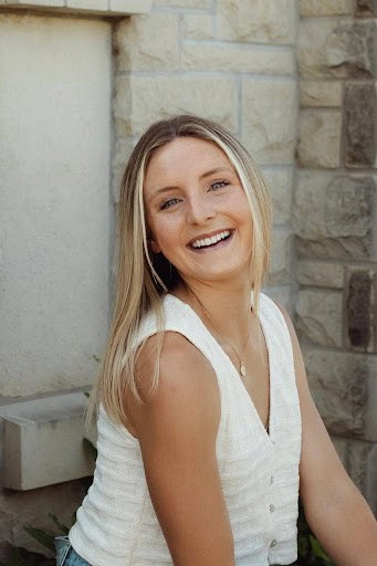 A young woman with straight, blonde hair and a sleeveless white top smiles while sitting in front of a stone wall, looking slightly over her shoulder—radiating confidence and positivity inspired by Unbound Recovery.