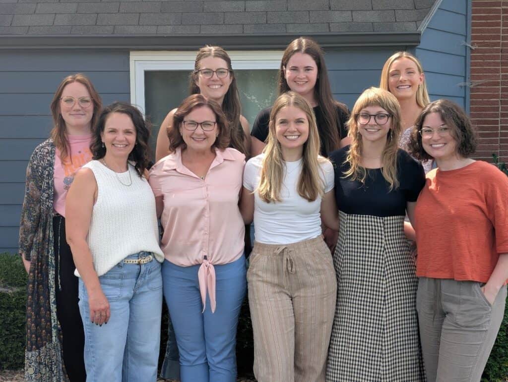 Nine women stand together outside in front of a blue house, smiling at the camera. Dressed in casual, summery outfits, they appear to be enjoying a sunny day while celebrating recent Unbound Recovery updates.