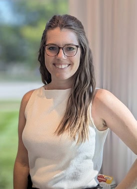 A woman with long brown hair and glasses smiles at the camera, celebrating National Nutrition Month in March. She wears a sleeveless white top, standing with one hand on her hip against a blurred green outdoor background.