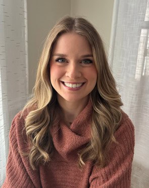 A woman with long, wavy blonde hair and a warm smile sits indoors by a window. She is wearing a textured, rust-colored sweater. Light filters through white curtains behind her, creating a cozy setting perfect for celebrating National Nutrition Month in March.
