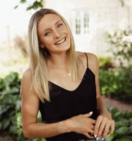 A young woman with long blonde hair smiles outdoors, wearing a black sleeveless top and a necklace, standing in front of greenery and a stone building, celebrating National Nutrition Month this March.