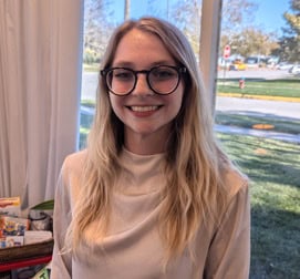 A young woman with long blonde hair and glasses smiles at the camera, celebrating National Nutrition Month. She is wearing a cream-colored top and stands indoors near a window, with trees, grass, and a street visible outside.