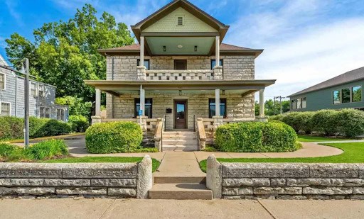 A two-story stone house with a wide front porch, double staircases, and neatly trimmed bushes offers a peaceful setting for healing, surrounded by a well-maintained lawn and stone fence under a blue sky.