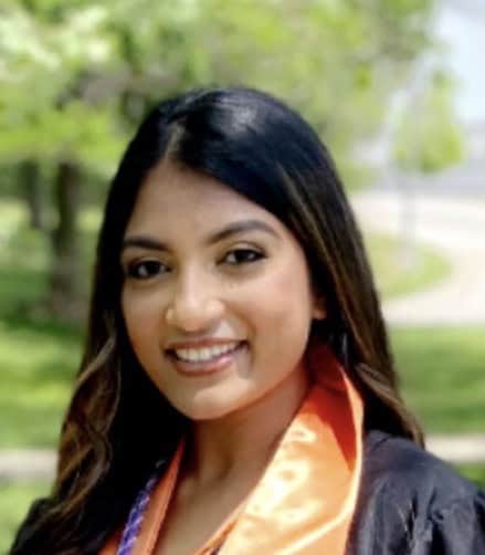A young woman wearing a graduation gown and an orange stole smiles outdoors, celebrating National Nutrition Month, with greenery and a pathway blurred in the background.