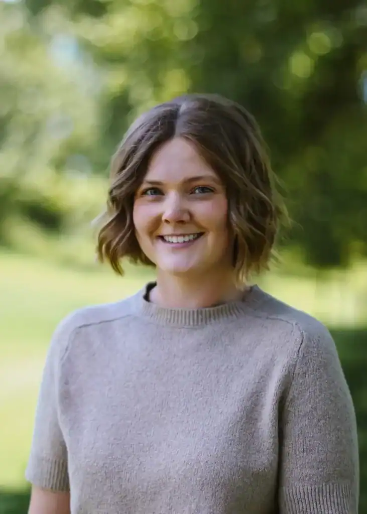 Lauren Ailslieger, a young woman with short, wavy brown hair, smiles at the camera while standing outdoors in a light beige, short-sleeve sweater, framed by a blurred background of green trees and grass.